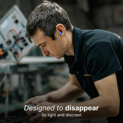 Photograph of a man in a workshop intently examining electronics, wearing a dark polo shirt and a bright blue earbud in his right ear against a background of industrial equipment