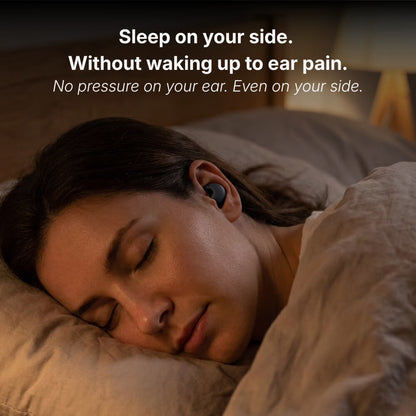 A woman sleeping comfortably on her side in a dimly lit bedroom, wearing a black wireless earbud, resting on a beige pillow, with warm light from a bedside lamp and overlay text reading Sleep on your side Without waking up to ear pain No pressure on your ear Even on your side
