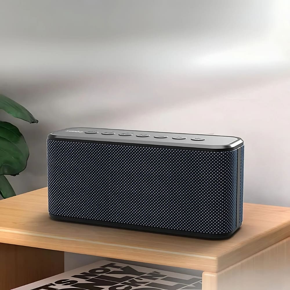 A black rectangular Bluetooth speaker featuring a dark blue woven mesh grille and silver control panel rests on a light wooden shelf with a green plant partially visible