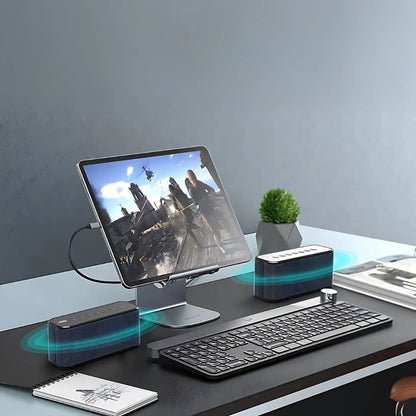 A modern desk setup displaying a tablet on a silver stand playing an action video game with a helicopter, accompanied by two black wireless speakers showing blue sound waves, a sleek black keyboard with a silver rotary dial, and a small green potted plant